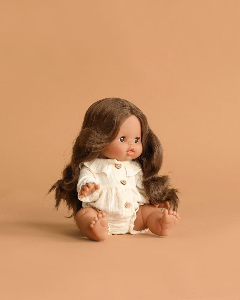 The Minikane Baby Doll (13") - Leopoldine with Clothing and Stroller Set features an anatomically correct doll with long brown hair, wearing a cream romper, posed on a matching light brown background facing slightly left.