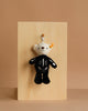 The Steiff Mr. Bones Teddy Bear Keychain, dressed in black skeleton-themed clothing, hangs from a silver hook on a light wooden board—a perfect Halloween accessory against the beige background.