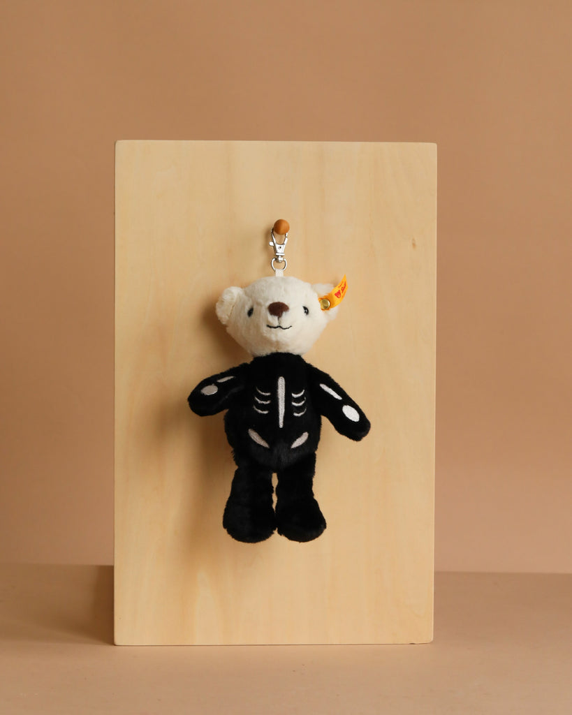 The Steiff Mr. Bones Teddy Bear Keychain, dressed in black skeleton-themed clothing, hangs from a silver hook on a light wooden board—a perfect Halloween accessory against the beige background.