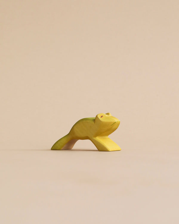 A Handmade Holzwald Jumping Frog sculpture in a crouching position on a plain, light beige background. The sculpture is painted in a vibrant yellow-green color, capturing a simplistic yet expressive.