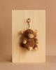 The Mon Ami Harold Highland Cow Keychain/Bag Charm, a plush brown and beige accessory, hangs from a wooden board against a beige background—ideal for adding a cute touch to your bag or keys.