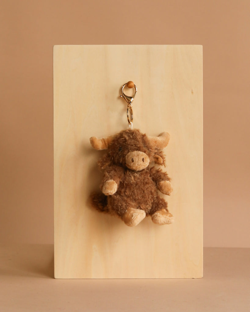 The Mon Ami Harold Highland Cow Keychain/Bag Charm, a plush brown and beige accessory, hangs from a wooden board against a beige background—ideal for adding a cute touch to your bag or keys.