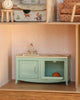 The Maileg Suitcasa Furnished Set includes a miniature dollhouse kitchen with mouse-sized light green sink cabinet, silver faucet, an orange oven item, and pink-and-white striped wallpaper accented with blue flower patterns.