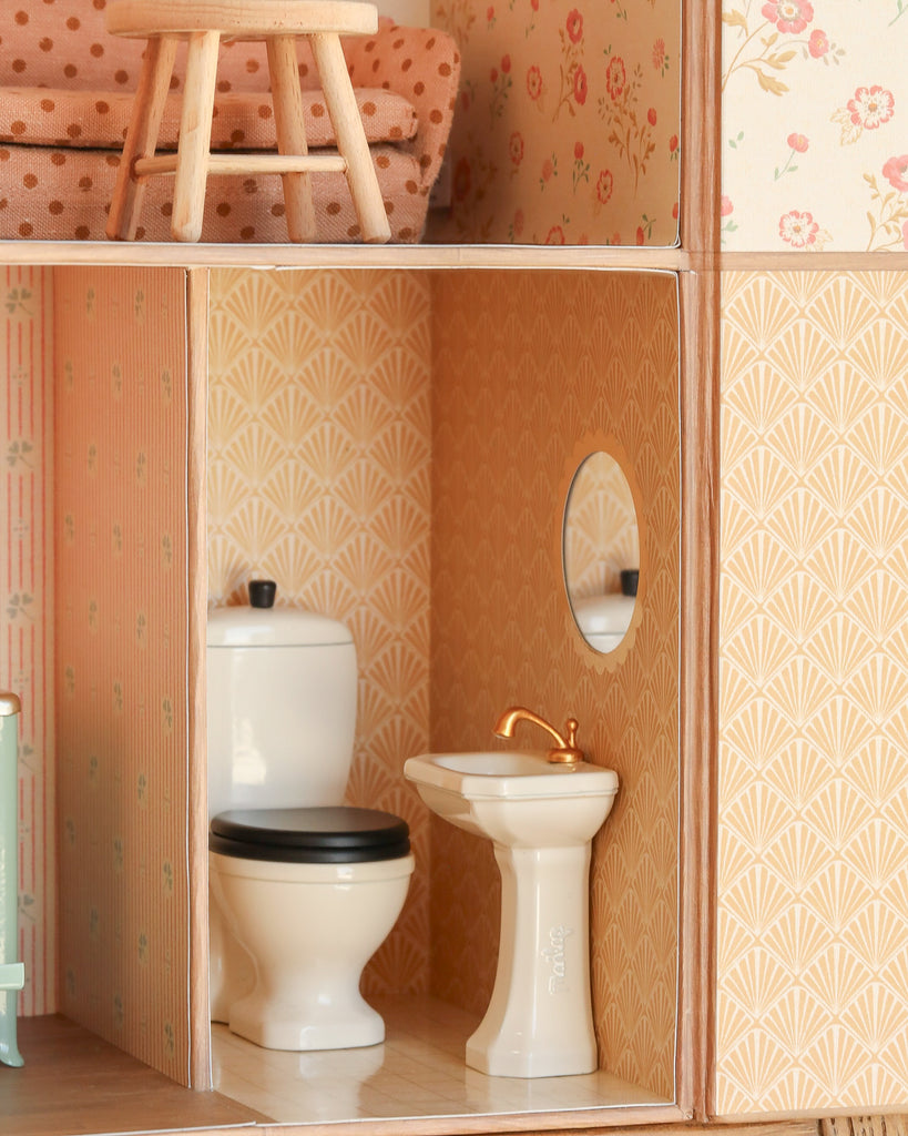 A Maileg Suitcasa Furnished Set bathroom features a white toilet, sink with gold faucet, round mirror, and beige patterned wallpaper. Above, part of a bedroom with Maileg mouse furniture and a polka-dot couch is visible.