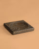 A square, black oak Mader Black Oak Spinning Plate with a circular indentation and natural wood grain patterns rests on a plain light brown background. Crafted to meet toy safety standards.