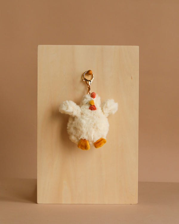 The Mon Ami - Hazel Hen Keychain / Bag Charm is a fluffy white chicken plush with an orange beak and feet on a gold keyring, ideal for adding cute flair to your bag or keys. Displayed on a light wood board with a beige background.