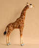 The Medium Giraffe Stuffed Animal, 34''—a hand-sewn plush toy with brown and white fur, a long neck, black tail tip, and gray hooves—stands upright against a beige background. FINAL SALE from HANSA animals.