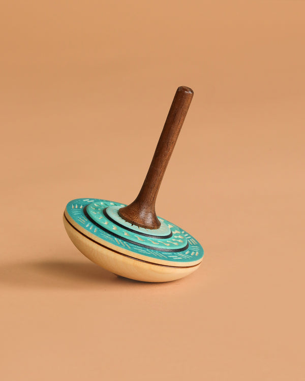 The Mader Bonbon Spinning Top - Light Blue is an Austrian wooden top with an embossed teal and beige base and a dark brown handle, photographed on a plain light brown background.