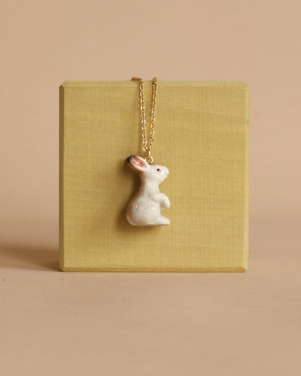 The White Rabbit Necklace features a 24k gold plated pendant shaped like a small white rabbit with pink ears, showcased against a light yellowish-brown square background. The minimalistic design and arrangement highlight the heirloom quality of the piece, set against a plain beige backdrop.