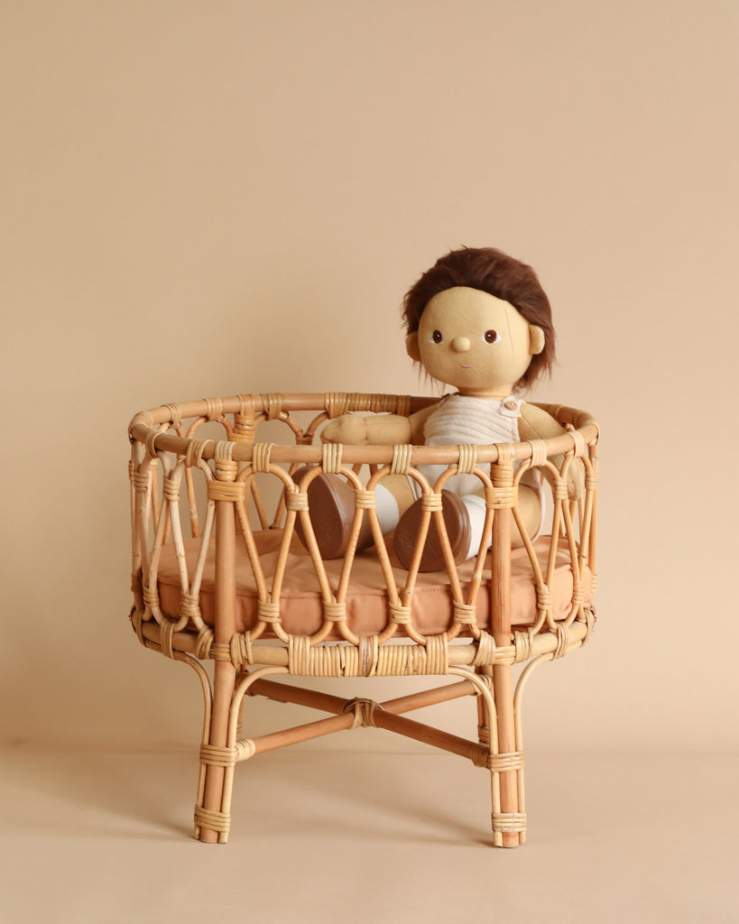 A Poppie Rattan Doll Crib + Duvet Set with brown hair seated in a small rattan peacock chair against a plain beige background. The doll is dressed in a white and blue striped outfit.