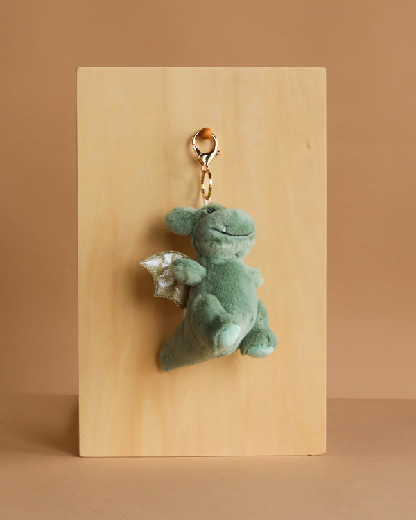 The Mon Ami - Dax Dragon Keychain / Bag Charm features a small green plush dragon with silver wings and a gold clasp, displayed on a light wooden board—ideal as an adorable plush charm or unique bag accessory.