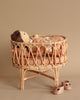 A Poppie Rattan Doll Crib + Duvet Set lies in a rattan crib with a plush toy by its side, next to a pair of baby shoes on a beige background.