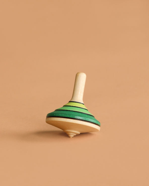The Mader Small Tango Spinning Top - Green, made of maple wood with green and yellow stripes, stands upright on a light brown surface against a plain beige background.
