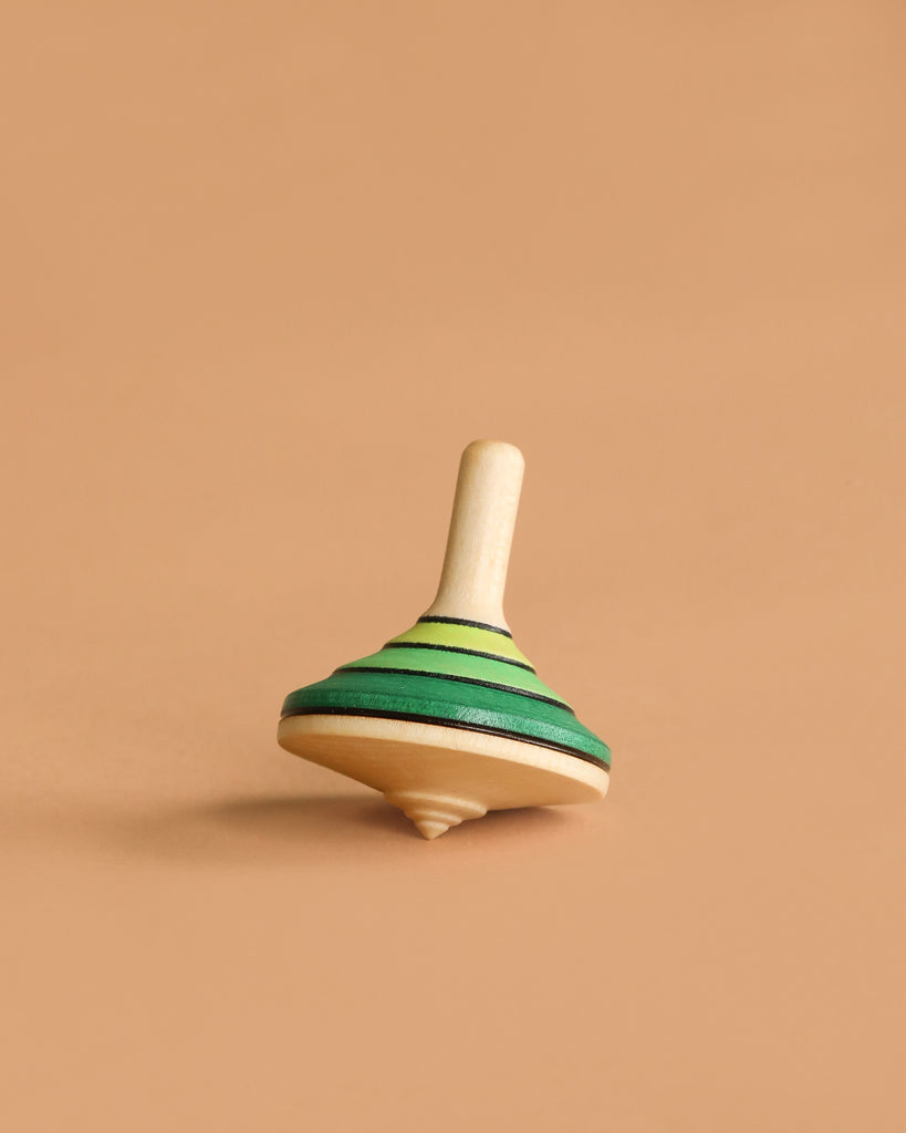 The Mader Small Tango Spinning Top - Green, made of maple wood with green and yellow stripes, stands upright on a light brown surface against a plain beige background.