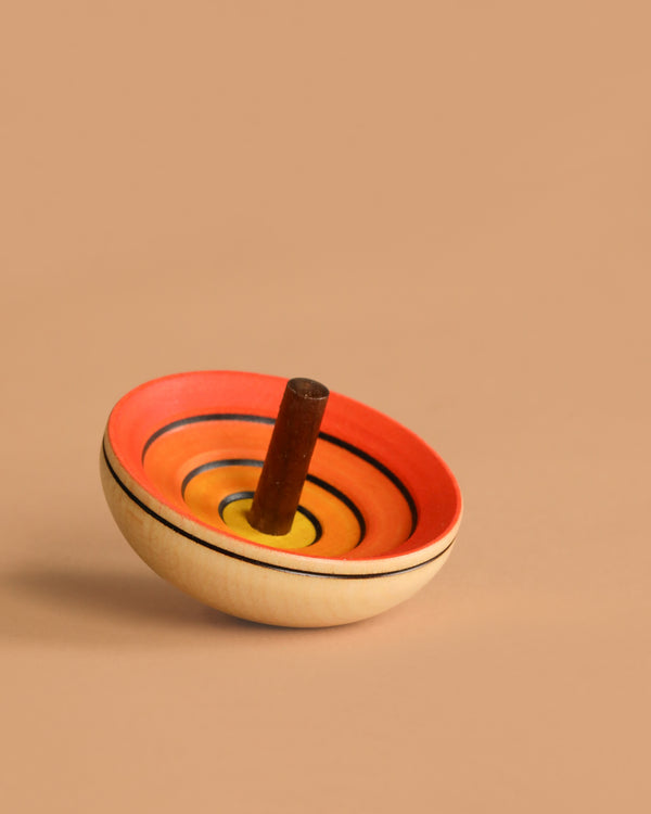 The Mader UFO Flora Spinning Top - Yellow, handcrafted from maple wood with ombre rings in red, orange, yellow, and black, rests on its side against a plain light brown background.