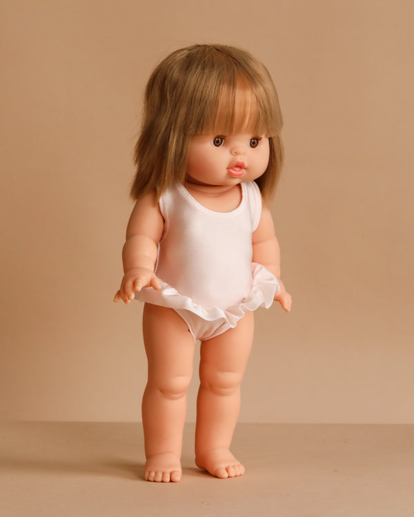 The Minikane Standing Doll (14") - Zoelie With Clothing features light skin, straight brown hair with bangs, and wears a white sleeveless leotard with a ruffled skirt. This anatomically correct doll is made in Spain.