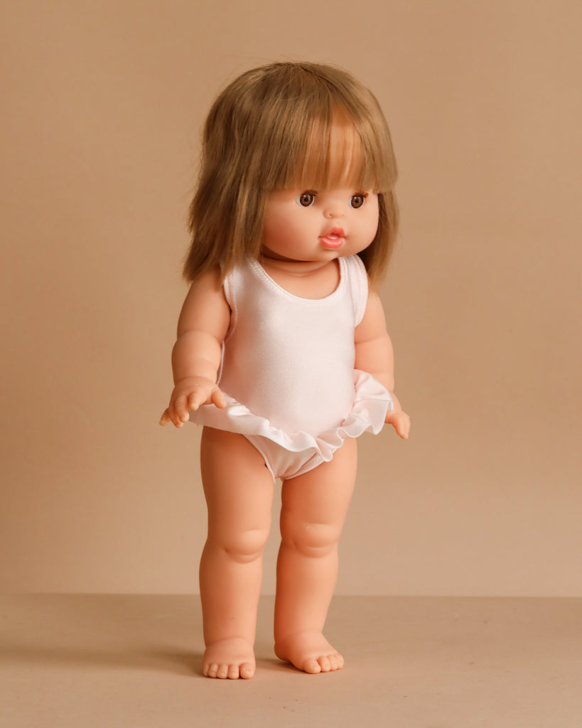 The Minikane Standing Doll (14") - Zoelie With Clothing features light skin, straight brown hair with bangs, and wears a white sleeveless leotard with a ruffled skirt. This anatomically correct doll is made in Spain.