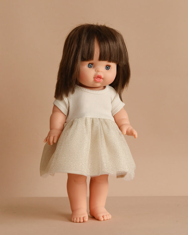 The Minikane Standing Doll (14") - Chléa With Clothing features straight brown hair, blue eyes, and a light complexion. She stands upright barefoot in a white short-sleeved dress with a sparkly skirt against a beige background.