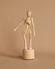 A "Wooden Lazy Bones Skeleton Pop Up" sculpture features a small skeleton figurine with simplified, jointed limbs standing on a cylindrical base, set against a beige background. The standing skeleton prominently displays a large wooden heart where its ribcage would be.