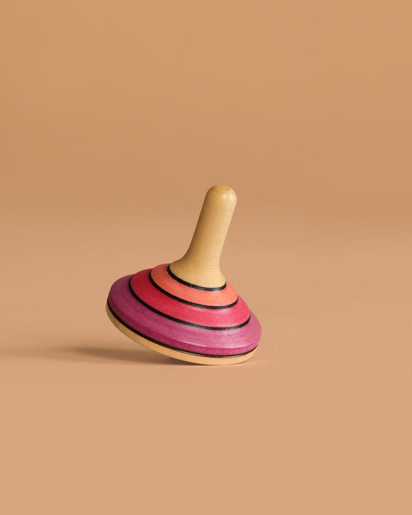 The Mader Small Tango Spinning Top - Pink, featuring vibrant pink, orange, and black stripes on maple wood, is balanced on its edge against a plain beige background.