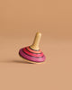 The Mader Small Tango Spinning Top - Pink, featuring vibrant pink, orange, and black stripes on maple wood, is balanced on its edge against a plain beige background.
