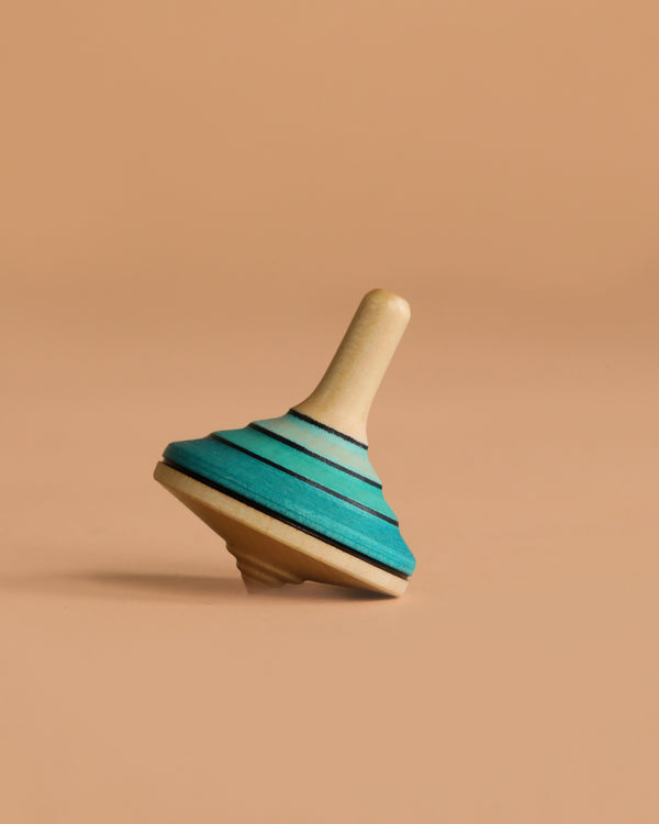The Mader Small Tango Spinning Top - Light Blue, made from maple wood, is slightly tilted on a plain beige background.