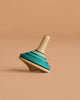 The Mader Small Tango Spinning Top - Light Blue, made from maple wood, is slightly tilted on a plain beige background.