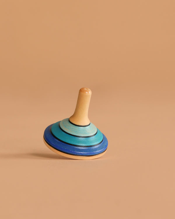 The Mader Small Tango Spinning Top - Dark Blue, crafted from Maple wood with blue and teal rings, stands balanced upright on a smooth light brown surface with a matching beige background.
