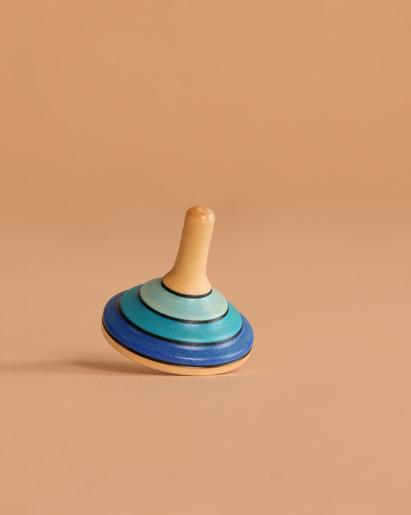 The Mader Small Tango Spinning Top - Dark Blue, crafted from Maple wood with blue and teal rings, stands balanced upright on a smooth light brown surface with a matching beige background.