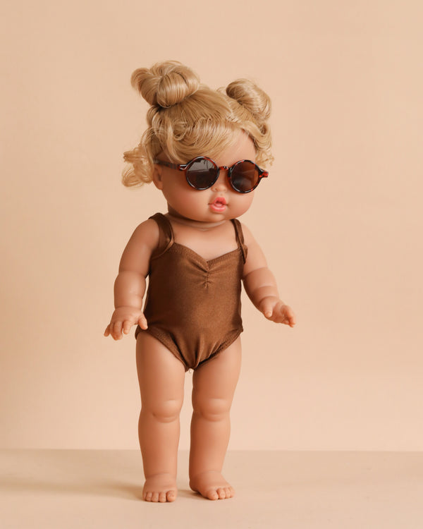 The Minikane Standing Doll (14") - Louisa with Clothing features blonde hair in two buns, dark sunglasses, a brown swimsuit, and stands barefoot against a beige background—an anatomically correct doll perfect for creative play.
