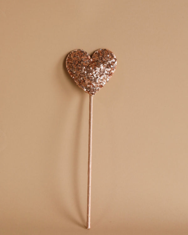 A Magic Wand - Pink Sequin Heart, adorned with glittery accents, stands against a plain beige background. This charming creation by Luciole et Petit Pois is attached to a thin stick and sparkles with whimsical elegance.