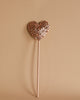 A Magic Wand - Pink Sequin Heart, adorned with glittery accents, stands against a plain beige background. This charming creation by Luciole et Petit Pois is attached to a thin stick and sparkles with whimsical elegance.