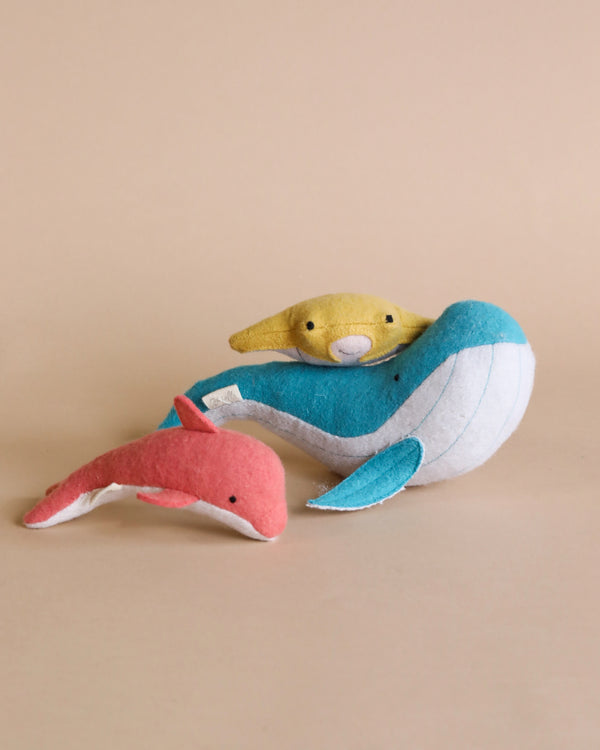 A plush toy sea otter lying back on a larger blue and white Olli Ella Holdie Folk Felt Ocean Animals whale, with a small orange and pink plush fish in the foreground, all set against a plain beige background.