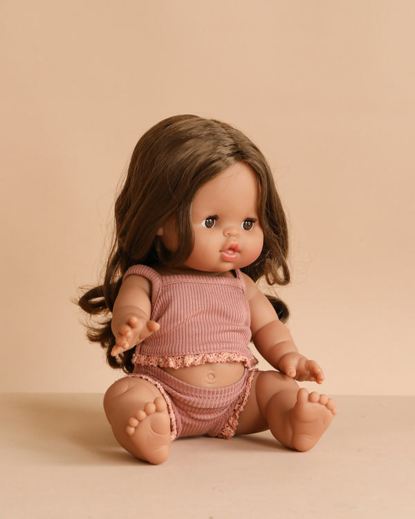 The Minikane Baby Doll (13") - Leopoldine With Clothing features brown hair and a soft expression, dressed in matching pink ribbed top and bottoms. This anatomically correct doll is lovingly made in Spain.