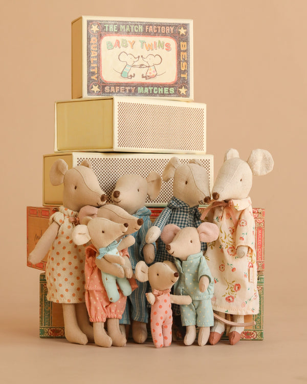 A group from the Maileg Mouse Family Collection, dressed in colorful pajamas and dresses, stand before a stack of Maileg matchbox decorations against a warm, neutral background.