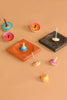 The Mader Cherry Wood Spinning Plate holds vibrant wooden spinning tops on a light brown surface, with some tops resting in the curved cherry plate and others scattered nearby, highlighting their colorful circular patterns.