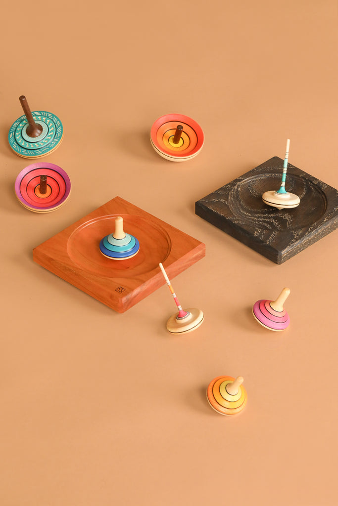 The Mader Cherry Wood Spinning Plate holds vibrant wooden spinning tops on a light brown surface, with some tops resting in the curved cherry plate and others scattered nearby, highlighting their colorful circular patterns.