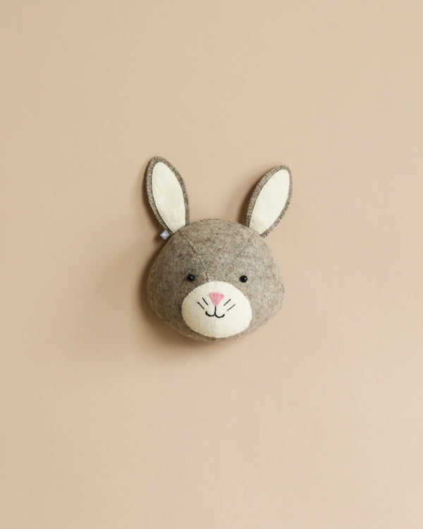 A Handcrafted Felt Bunny Wall Decor - Mini head mounted on a plain beige background, featuring soft gray organic wool fur, prominent white inner ears, and a cute, stitched face.