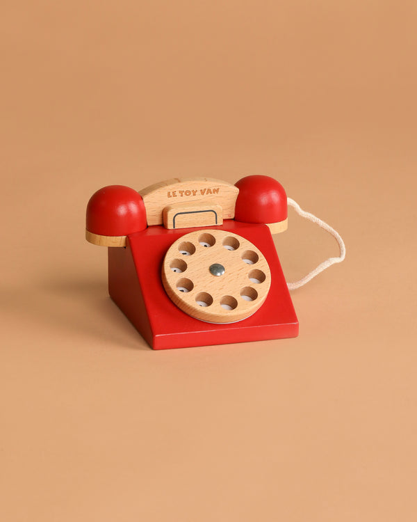 The Vintage Wooden Telephone is a red wooden toy phone with a natural wood dial and handset, connected by a short white cord, set against a plain beige background.