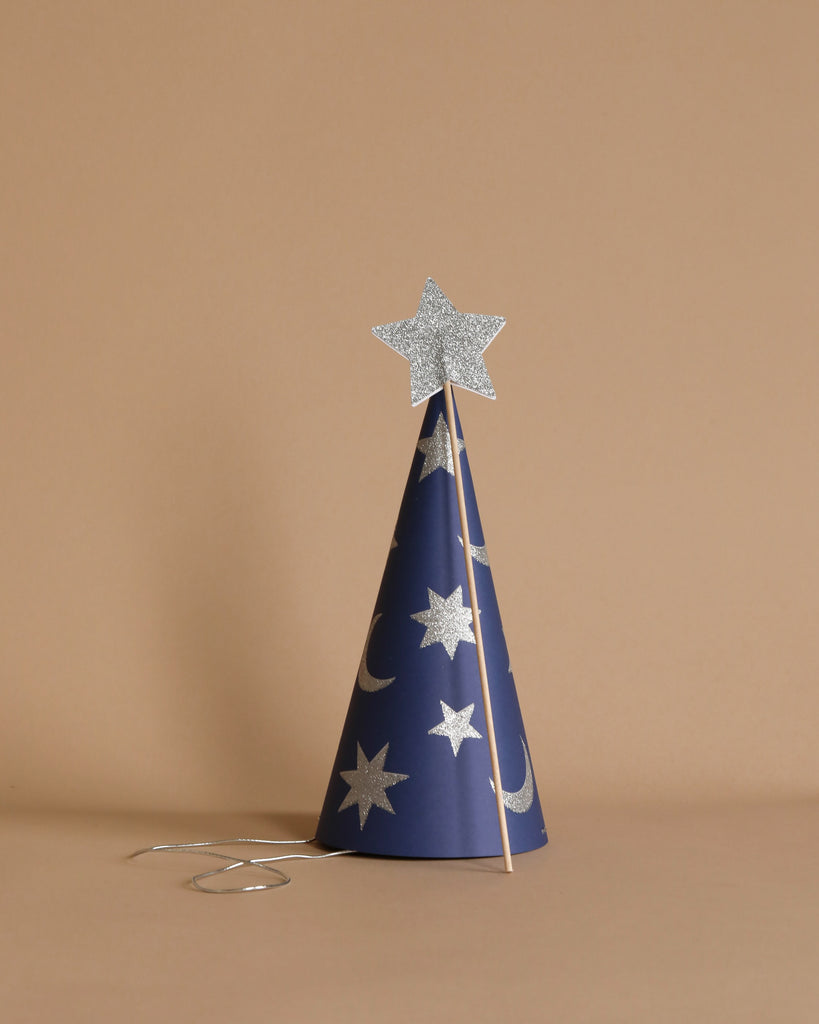 The Meri Meri Wizard Party Hat & Wand set features a dark blue, cone-shaped party hat adorned with silver stars and crescent moons, standing upright against a beige background. A shiny silver star on a stick, resembling a magic wand, leans gracefully against the hat, with a slender silver string extending from its base. This item is available as part of the final sale collection.