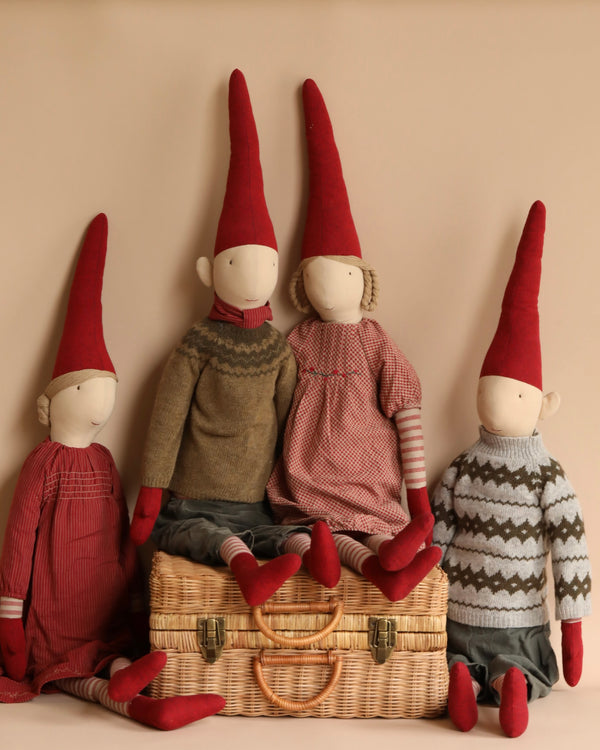 Four Maileg Christmas Mega Pixy dolls with pointed red hats, designed as Christmas decorations, seated on a wicker basket against a neutral background. Each doll is uniquely dressed in cozy sweaters and pants made from exclusive materials.