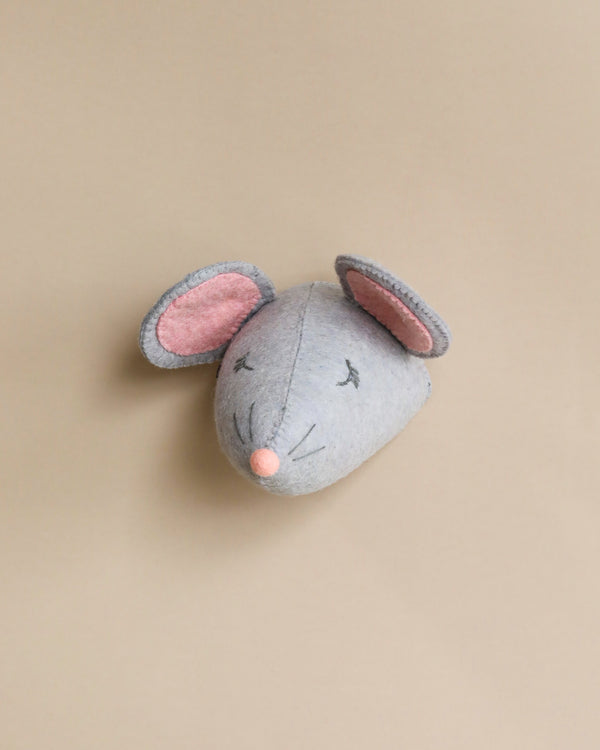 A Handcrafted Felt Mouse Wall Decor - Mini, handcrafted from organic wool with grey fabric, pink inner ears, closed eyes, and a stitched nose and whiskers, set against a plain beige background.