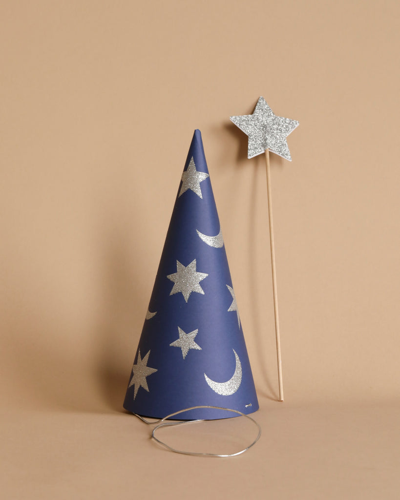 A blue, sparkly Meri Meri Wizard party hat adorned with silver stars and crescent moons stands upright on a beige surface. Beside it is a matching magic wand featuring a silver star on a wooden stick. A thin elastic strap at the base of the hat indicates where it can be worn.