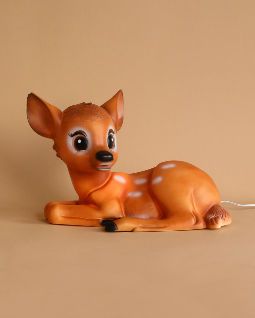 A photograph showcasing the Resin Deer Lamp, designed to resemble a baby deer, or fawn, in a lying-down position. The lamp features a light brown body adorned with white spots, expressive large eyes, and alert ears. Set against a plain beige background, the image also reveals a power cord near the hind legs of this charming LED night light, making it an ideal addition to nursery decor.