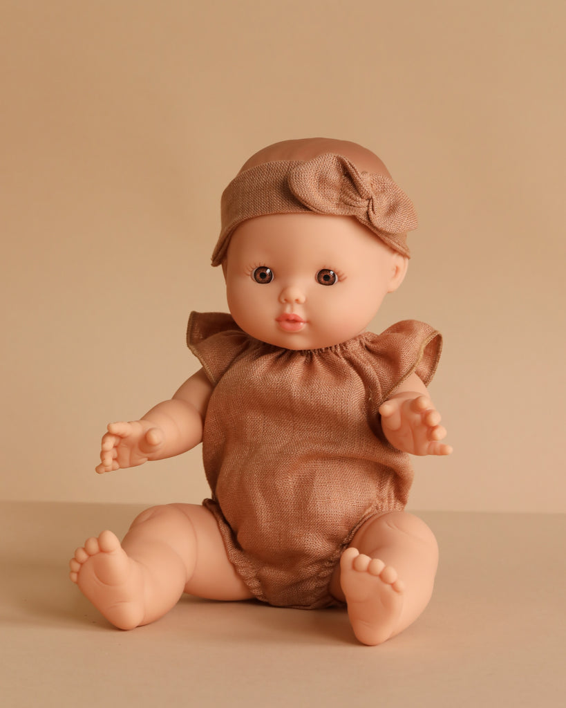 The Minikane Doll (13") - Faustine with Clothing, made in Spain from phthalate-free vinyl, has light skin and wears a beige headband with bow and matching romper while sitting on a beige surface against a beige background.