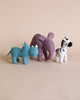 Three Olli Ella Holdie Folk Felt Safari Animals, a blue rhinoceros, a purple elephant, and a white and black zebra, standing in a line on a light beige background, perfect for imaginative play.