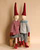 Two decorative Maileg Christmas Mega Pixy dolls seated on a wicker basket, one wearing a striped red and white shirt and the other in a grey and blue sweater, both with long red hats.