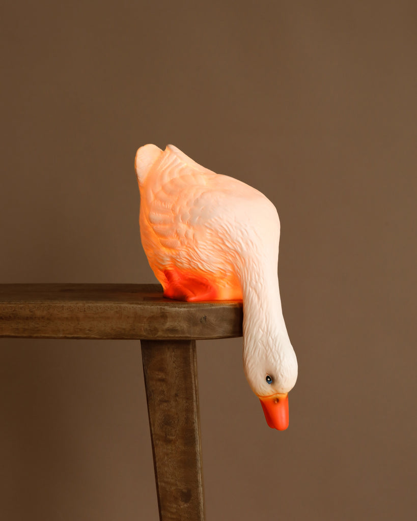 The Duck Looking Down Lamp, hand painted in white and orange, perches on a wooden table with its head leaning over the edge—an adorable accent for children's room decor against a simple brown backdrop.