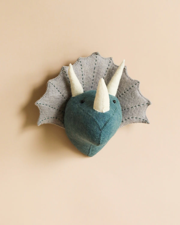 A handcrafted Felt Triceratops Wall Decor - Mini with two white horns and webbed frill, mounted on a beige wall. The dinosaur is in blue and gray colors.