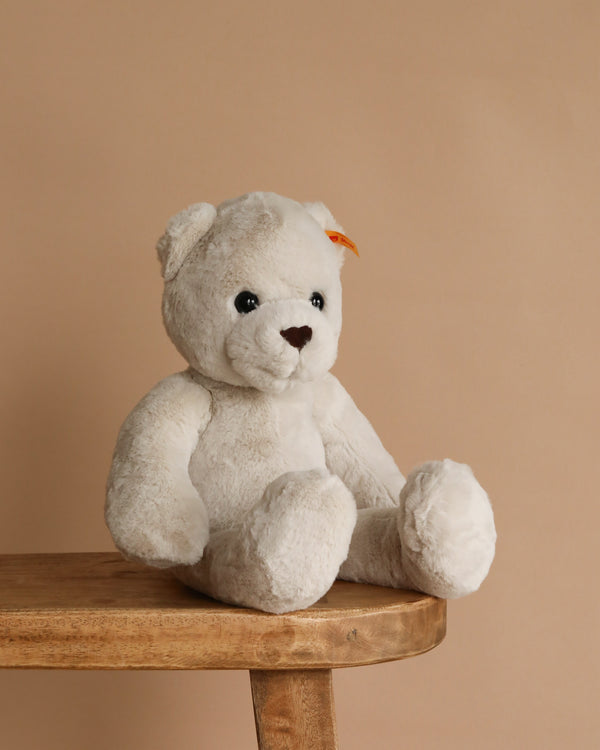 The Steiff Robby Teddy Bear 17" is a fluffy white plush toy with black eyes, a brown nose, and a small orange tag on its left ear, pictured sitting on a wooden table against a beige background.